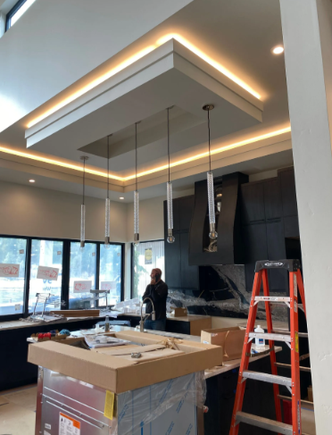 Partially finished modern kitchen with ceiling lights, ladder, and worker in front of windows