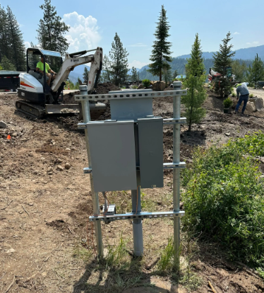 Utility boxes on metal frame beside a dirt road, with excavator and worker in a forested area.