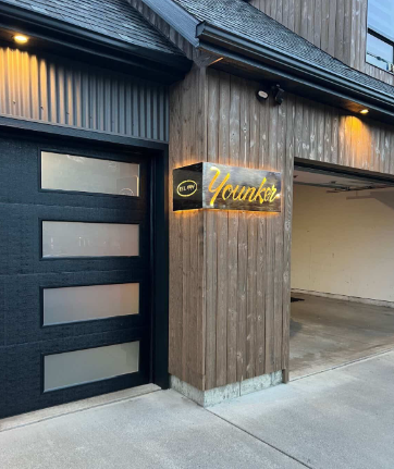 Modern storefront with a black garage-style door, wood siding, and a lit “Yomiko” sign.