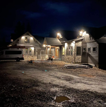 Nighttime construction site with two lit houses, a van, and gravel ground under a dark sky