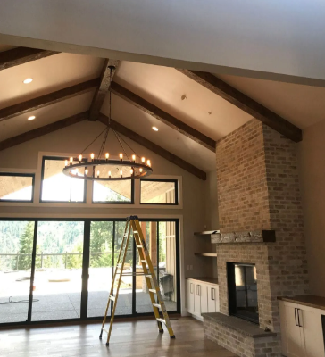 Living room with vaulted wood-beam ceiling, stone fireplace, chandelier, and a ladder by large windows