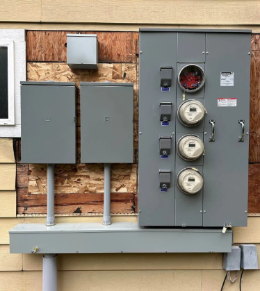 Gray electrical meter panel and utility boxes mounted on an exterior wooden wall.