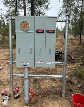 Gray electrical boxes on a metal frame beside a wooded path, with tools and a red bucket nearby