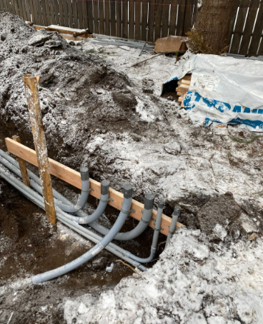 Trenched yard with gray pipes laid in snow and dirt, beside a wooden fence and plastic sheeting.