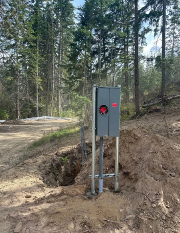 Gray electrical box on posts in a forest clearing beside a dirt trench and trees
