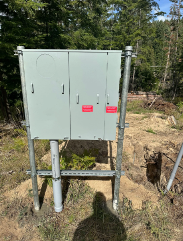 Gray utility cabinet on metal posts beside a dirt trench in a wooded area.