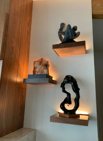 Three abstract sculptures on lit wall shelves against a white wall and wood paneling.