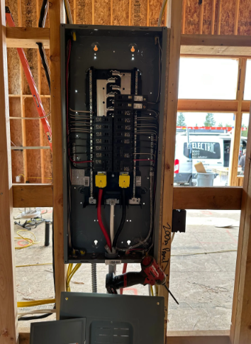 Electrical breaker panel installed in a wall opening at a construction site, with exposed wiring and studs