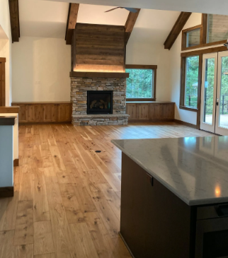 Open living room with wood floors, stone fireplace, exposed beams, and large windows, viewed from a kitchen island