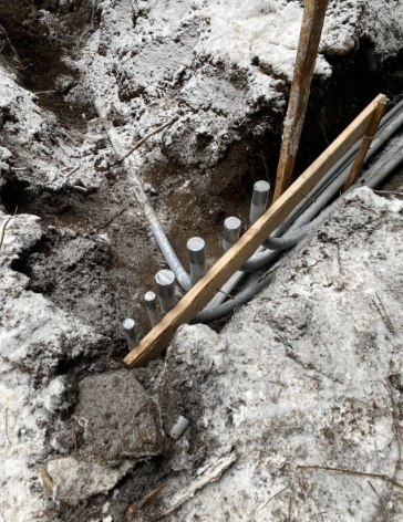 Rebar and wooden formwork in a muddy excavation trench, with concrete being poured into a narrow gap