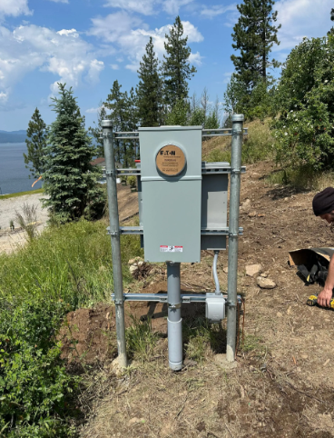 Gray utility box on metal frame near a lakeside trail, with trees and mountains in the background.