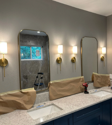 Bathroom vanity with two sinks, gray walls, gold sconces, and mirrors under renovation, with items covered in paper.