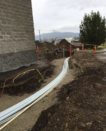 Construction trench with white cables running beside a building, orange cones, and dirt piles under cloudy sky