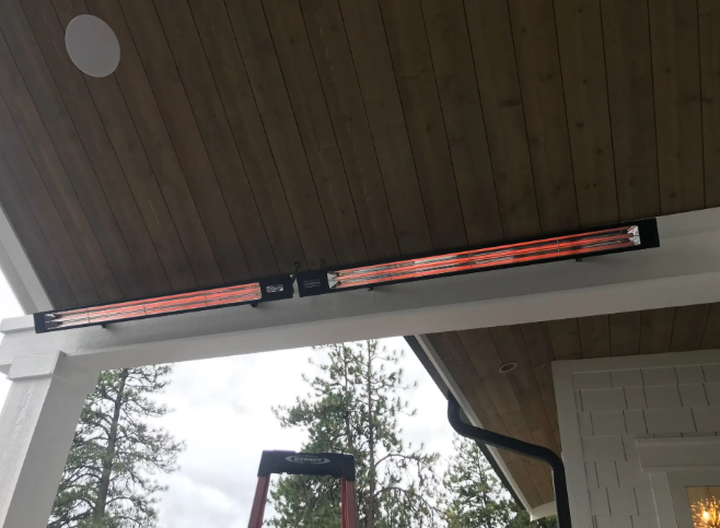 Under a porch roof, two red patio heaters are mounted above a white beam with trees beyond.