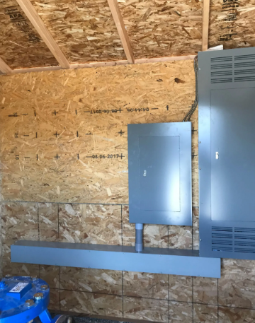 Electrical panels mounted on an OSB wall inside an unfinished building, with exposed framing overhead.