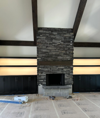 Modern living room with a stone fireplace, dark wood beams, and built-in cabinets under construction