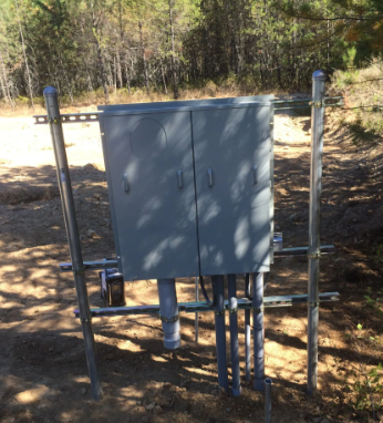 Gray utility box with pipes mounted on a metal frame outdoors near a wooded dirt road