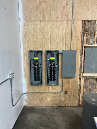 Two electrical panels mounted on plywood wall beside a gray breaker box in an unfinished room
