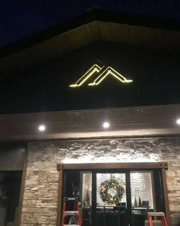 Nighttime storefront with a glowing mountain logo, stone facade, glass doors, and a holiday wreath inside.
