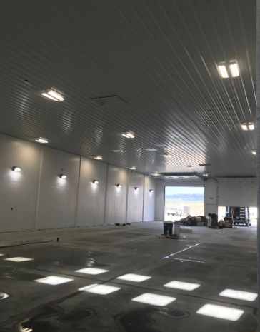 Large empty industrial hall with white walls, ceiling lights, and sunlight squares on the floor, tools at far end