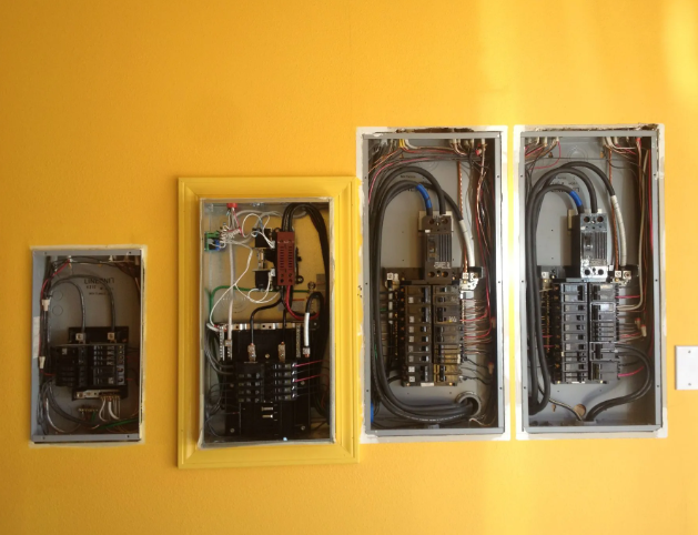 Four open electrical panels with wiring and components mounted on a yellow wall.
