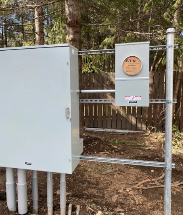 Utility boxes and electrical panel on a fenced dirt site in a wooded area