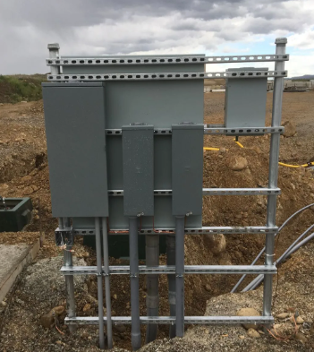 Electrical control boxes and conduit mounted on a metal frame in an outdoor gravel area.