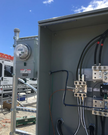 Open electrical control cabinet with meter, breakers, and wiring outdoors under blue sky