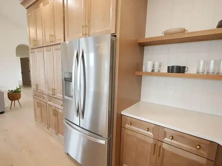 Stainless steel refrigerator in a light wood kitchen with white countertops and open shelving.