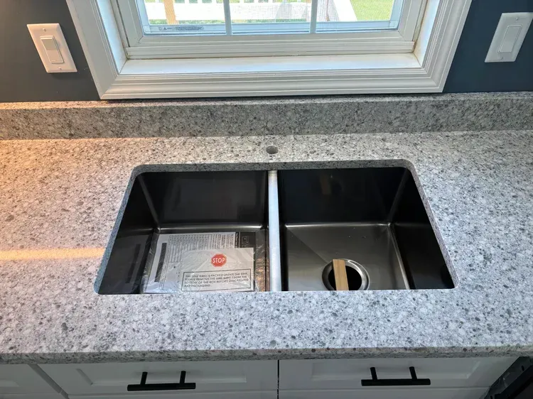 A kitchen sink with dual basins, installed in a speckled gray countertop, beneath a window.