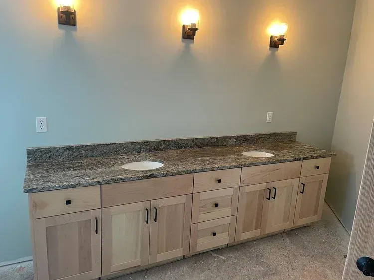 Bathroom vanity with granite countertop, two sinks, light wood cabinets, and three sconce lights on a teal wall.