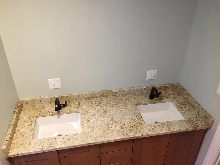 Double bathroom vanity with brown cabinets, light-colored granite countertop, white sinks, and black faucets, against a light green wall.
