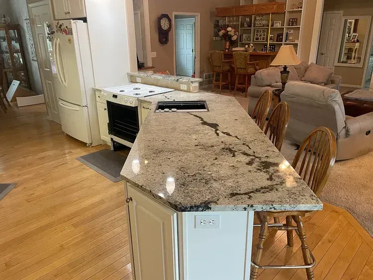 Kitchen island with granite countertop, stovetop, and seating. Wooden floors, refrigerator, and living room visible.