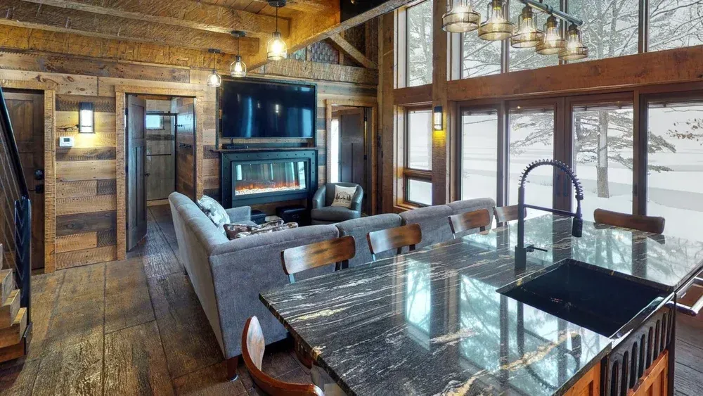 Rustic cabin interior with fireplace, kitchen island, large windows overlooking snowy landscape.