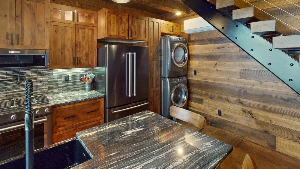 Rustic kitchen with wood cabinets, stainless steel appliances, and a stacked washer and dryer next to a staircase.