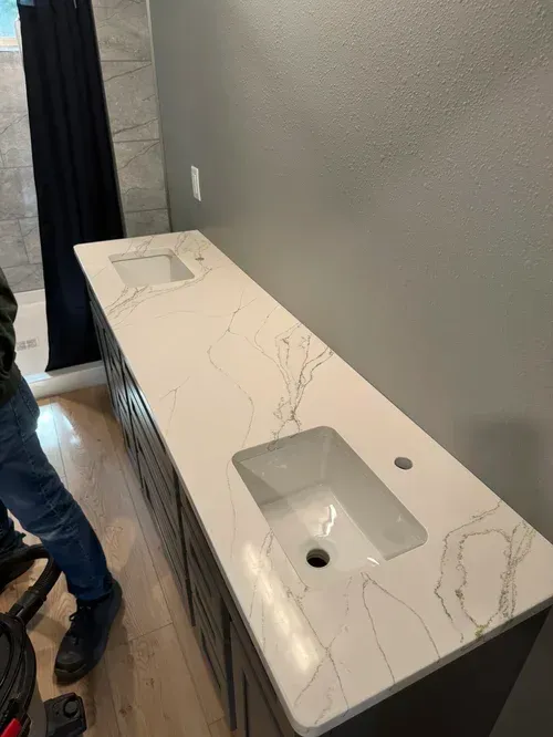 Bathroom vanity with white quartz countertop and two sinks, with person vacuuming. Gray walls.
