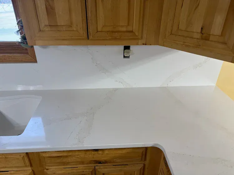 Kitchen with light-colored countertops and backsplash, beneath wooden cabinets. An electrical outlet is visible.