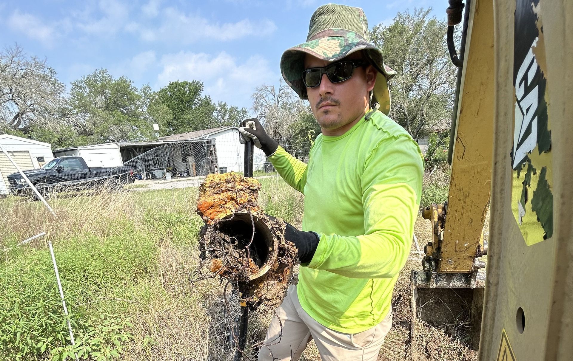 Worker in neon green shirt holding a muddy tool outdoors near a fence and trees.