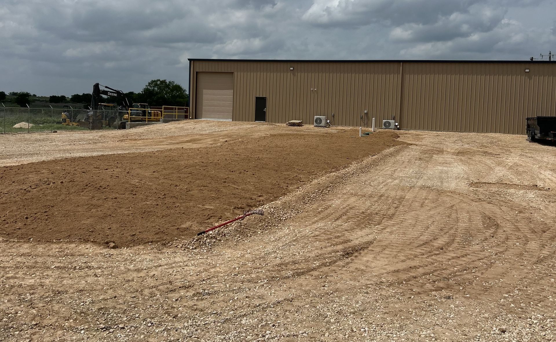 Dirt construction site with a large gravel mound and a tan metal building under cloudy skies.