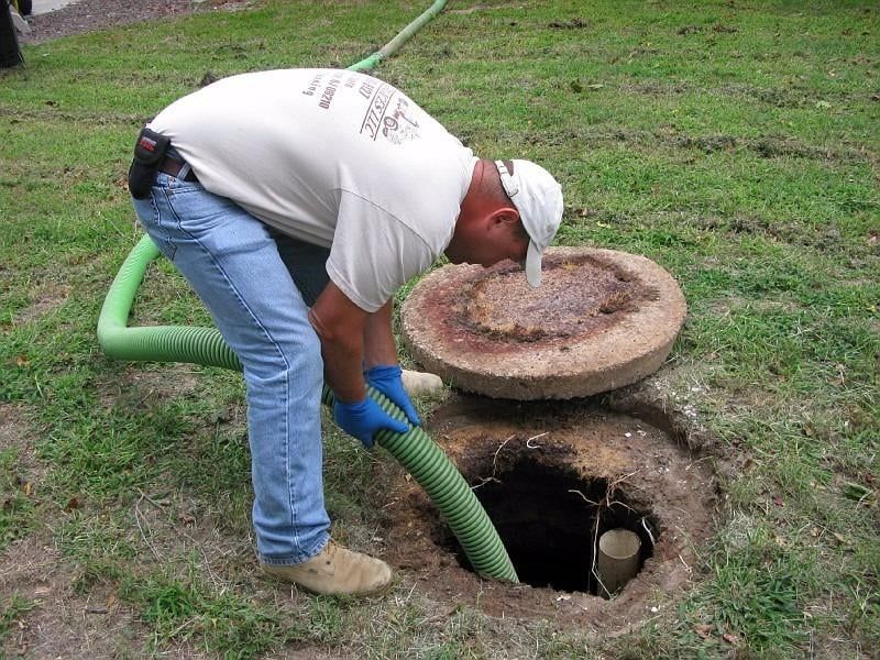 Worker in blue gloves clearing a clogged outdoor drain with a green hose.