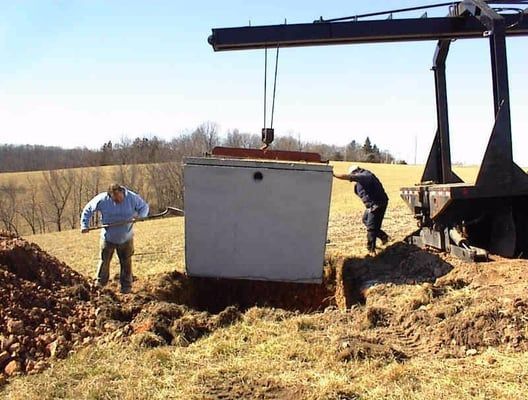 Workers lift a white concrete tank from a dirt trench with a crane in a rural field.