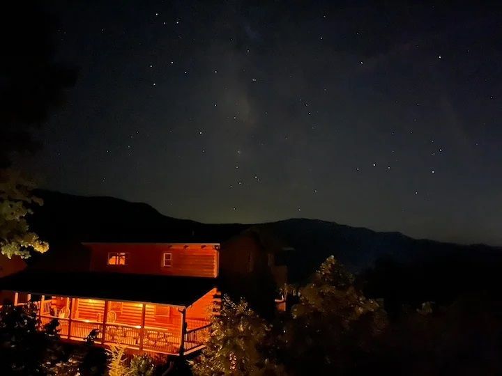 A cabin is lit up at night with a starry sky in the background.