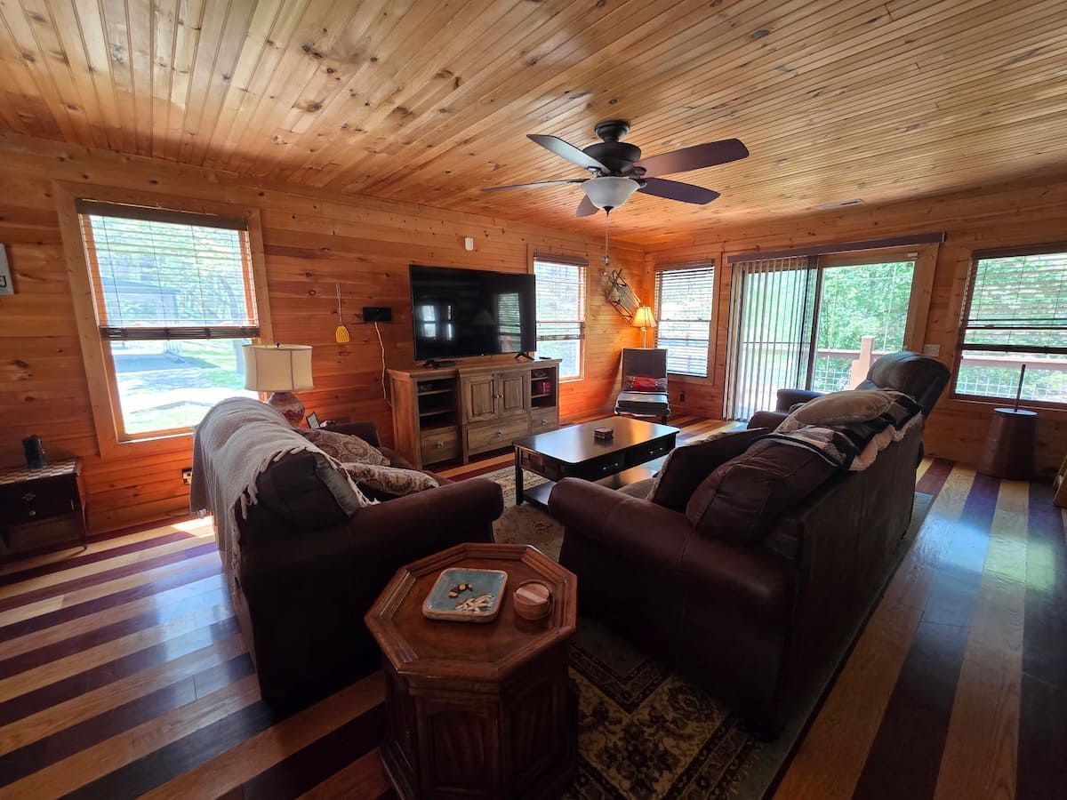 Cozy wood-paneled living room with sofas, TV, fireplace, and large windows overlooking greenery