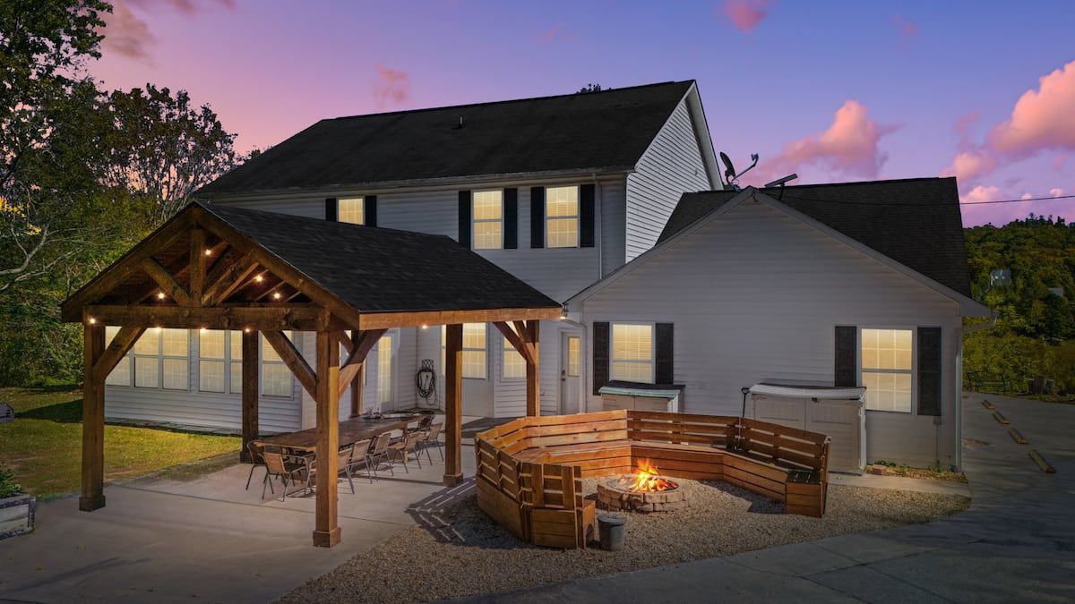 Two-story house at dusk with a wooden pergola, lit windows, and a fenced patio under a pink sky