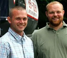 Two men smiling in front of a helicopter. One in plaid shirt, other in green polo.