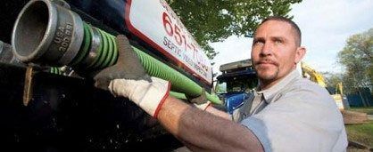 Man in work gloves attaching a green hose to a dark tank.