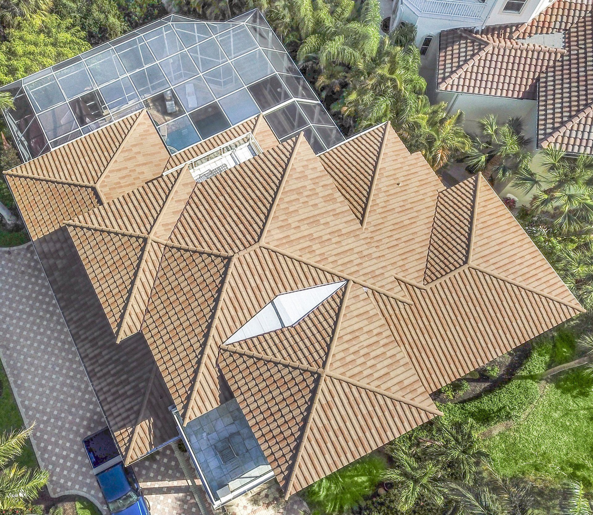Aerial view of a complex, multi-gabled tan shingle roof with a glass-covered patio section and surrounding greenery.