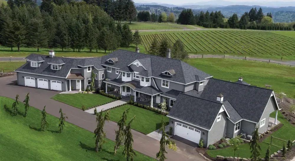 An aerial view of a large, gray, multi-roofed suburban house with a paved driveway and an adjacent vineyard.