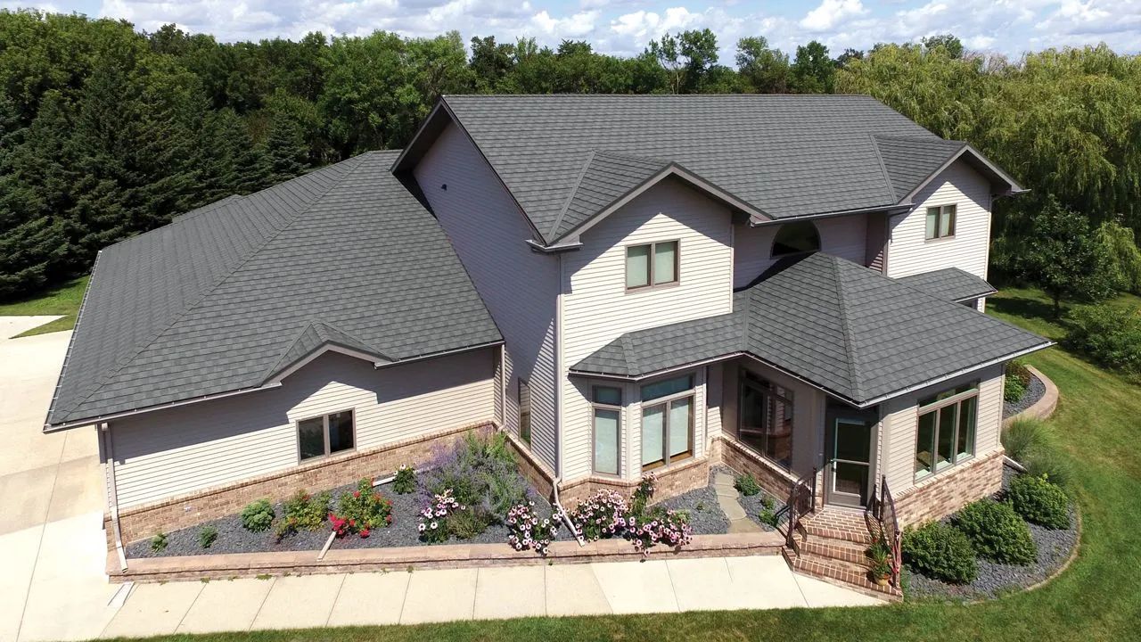 An aerial view of a two-story beige house with a grey roof, stone foundation, and landscaped flower beds.