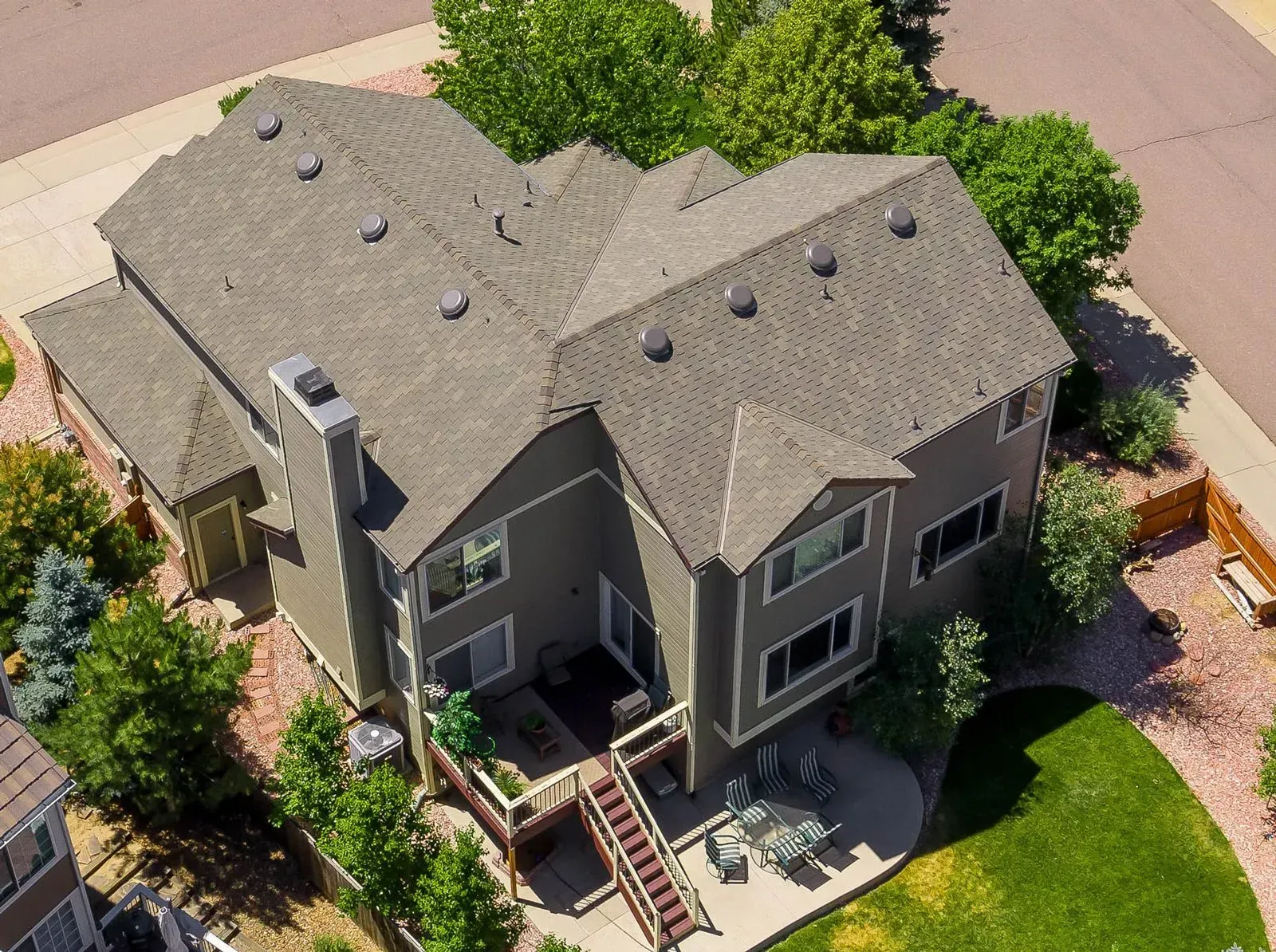 Aerial view of a two-story house with a grey roof, dark siding, a wooden deck, and a patio in a suburban neighborhood.